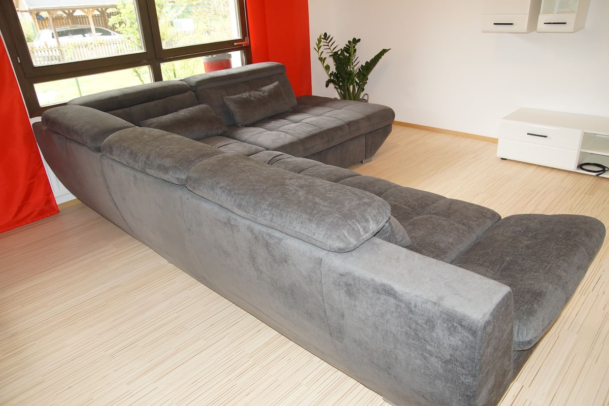 A modern living room with a grey sectional sofa, a white storage unit, and a large window with a view of a fence and greenery.
