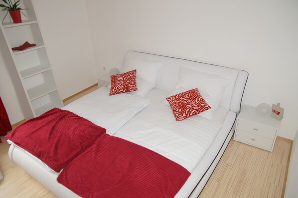 Una cama doble con ropa de cama blanca y dos almohadas rojas estampadas, un plaid rojo, una mesita de noche blanca con una lámpara, y una pequeña estantería con una planta en maceta y un par de zapatos.