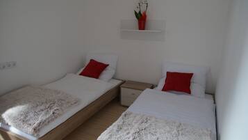 A bedroom with two single beds, each with a white mattress and red pillows, a wooden nightstand, and a small shelf with a plant.