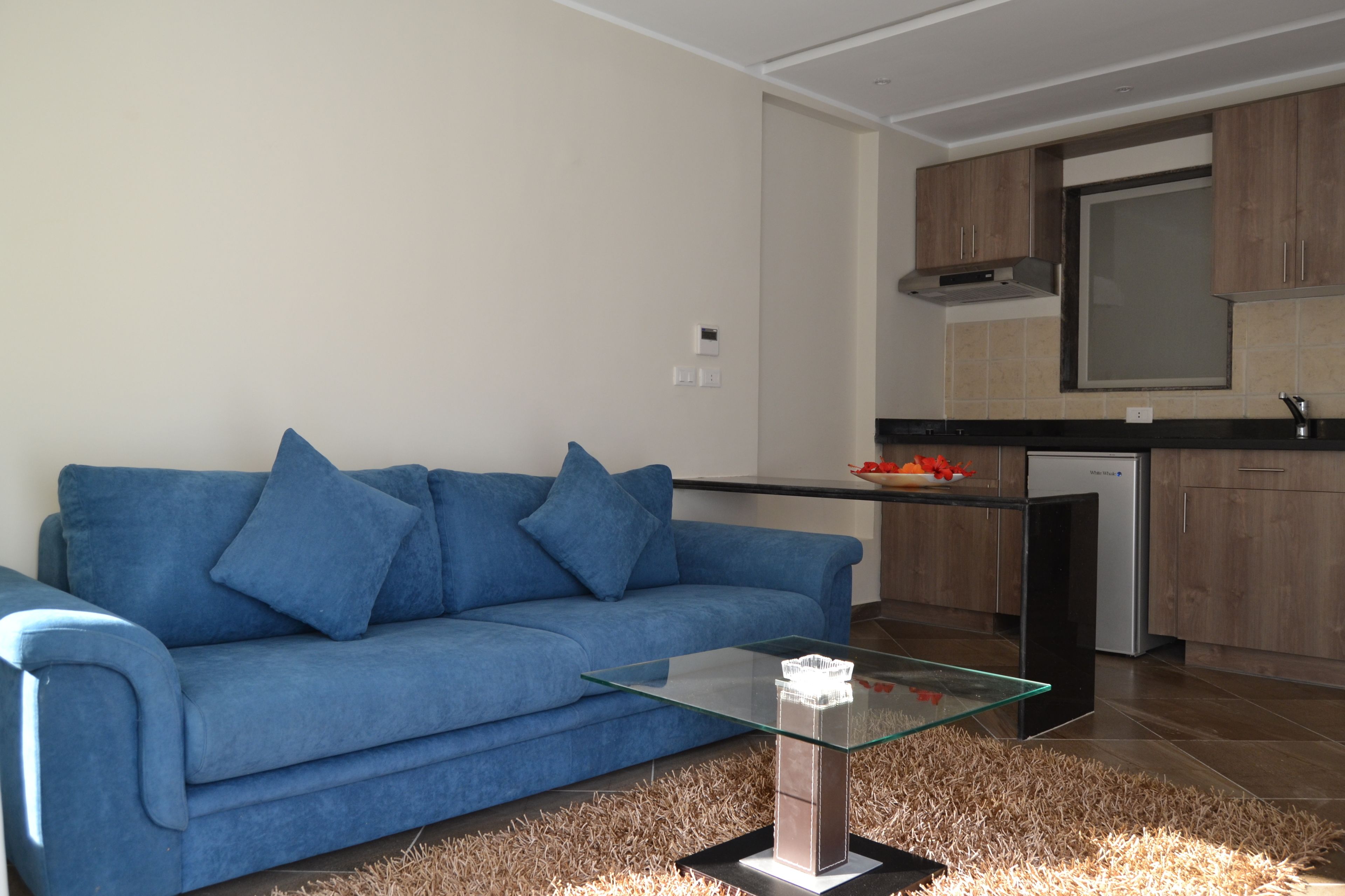 A modern living room with a blue sofa, glass coffee table, and a kitchen area with wooden cabinets and a built-in oven.