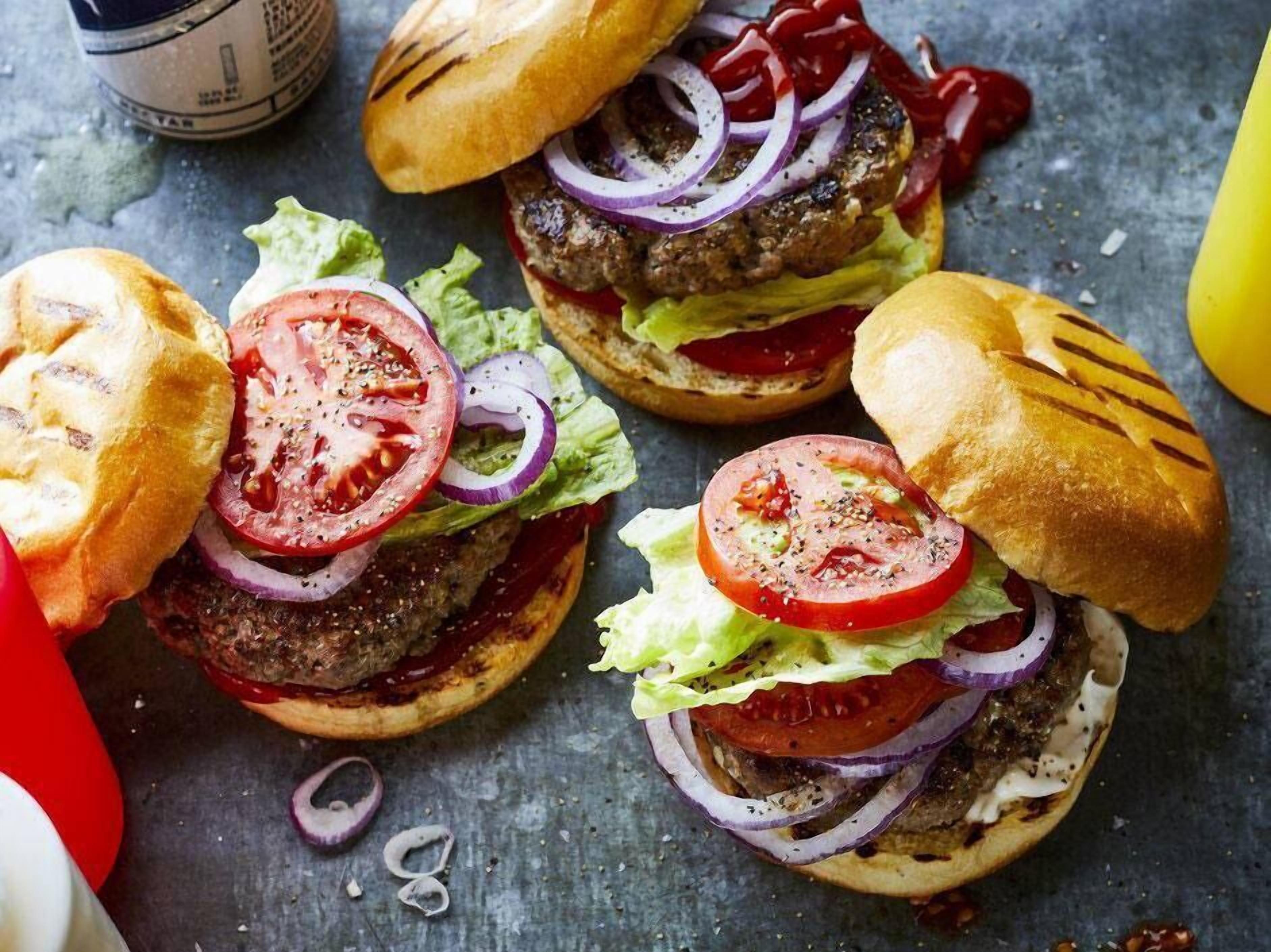 A close-up of three burgers with lettuce, tomato, and red onion on a textured surface.
