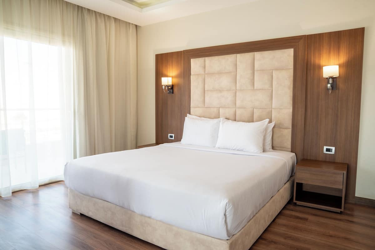 A large bed with a wooden headboard and two bedside lamps, a wooden nightstand, and a large window with curtains.