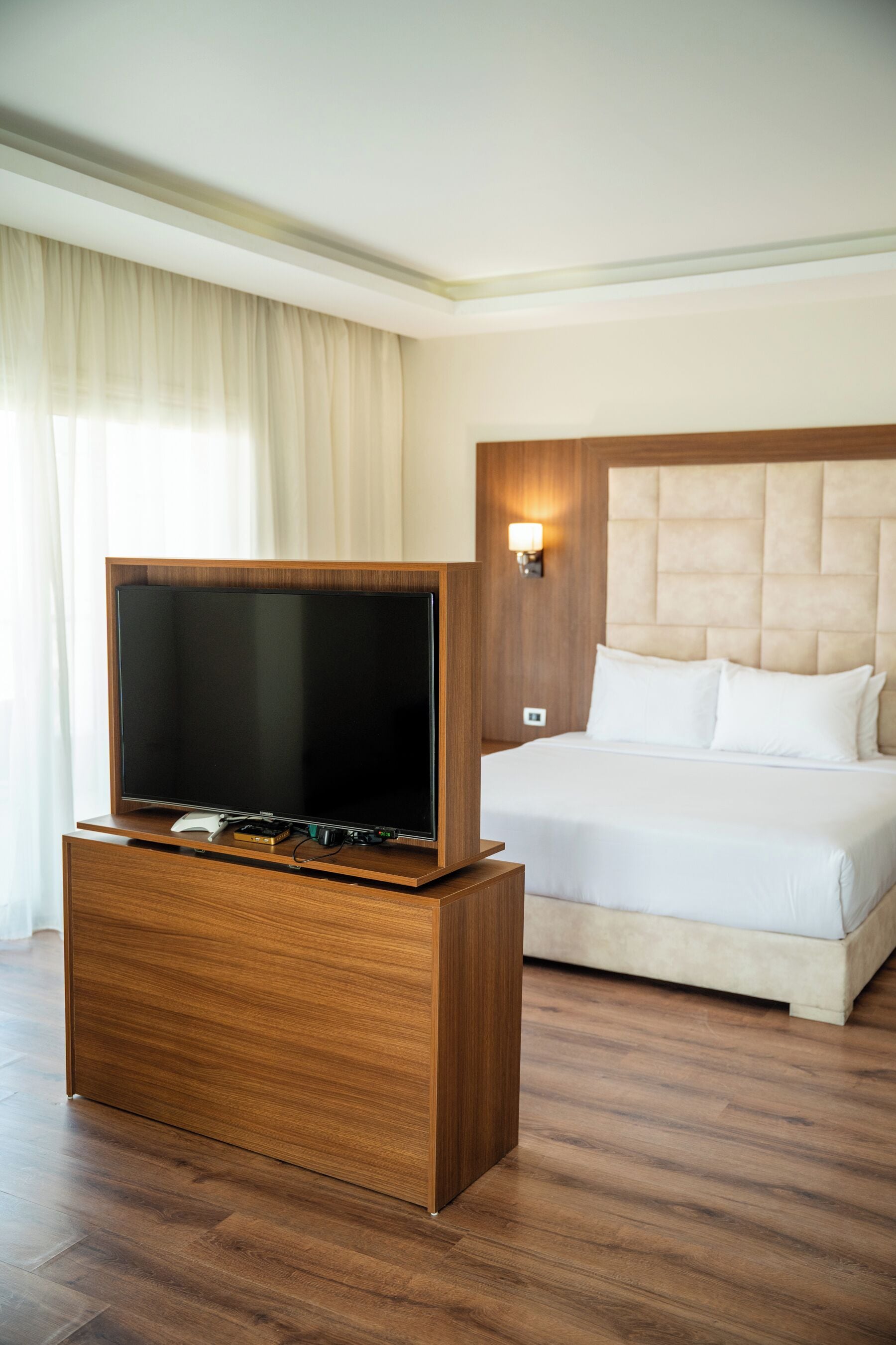 A hotel room with a wooden TV stand, a bed with a tufted headboard, and a wall-mounted light.