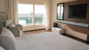 Habitación de hotel con una cama grande, un sofá, un televisor montado en la pared y un ventanal con vistas a la vegetación y un paisaje distante.