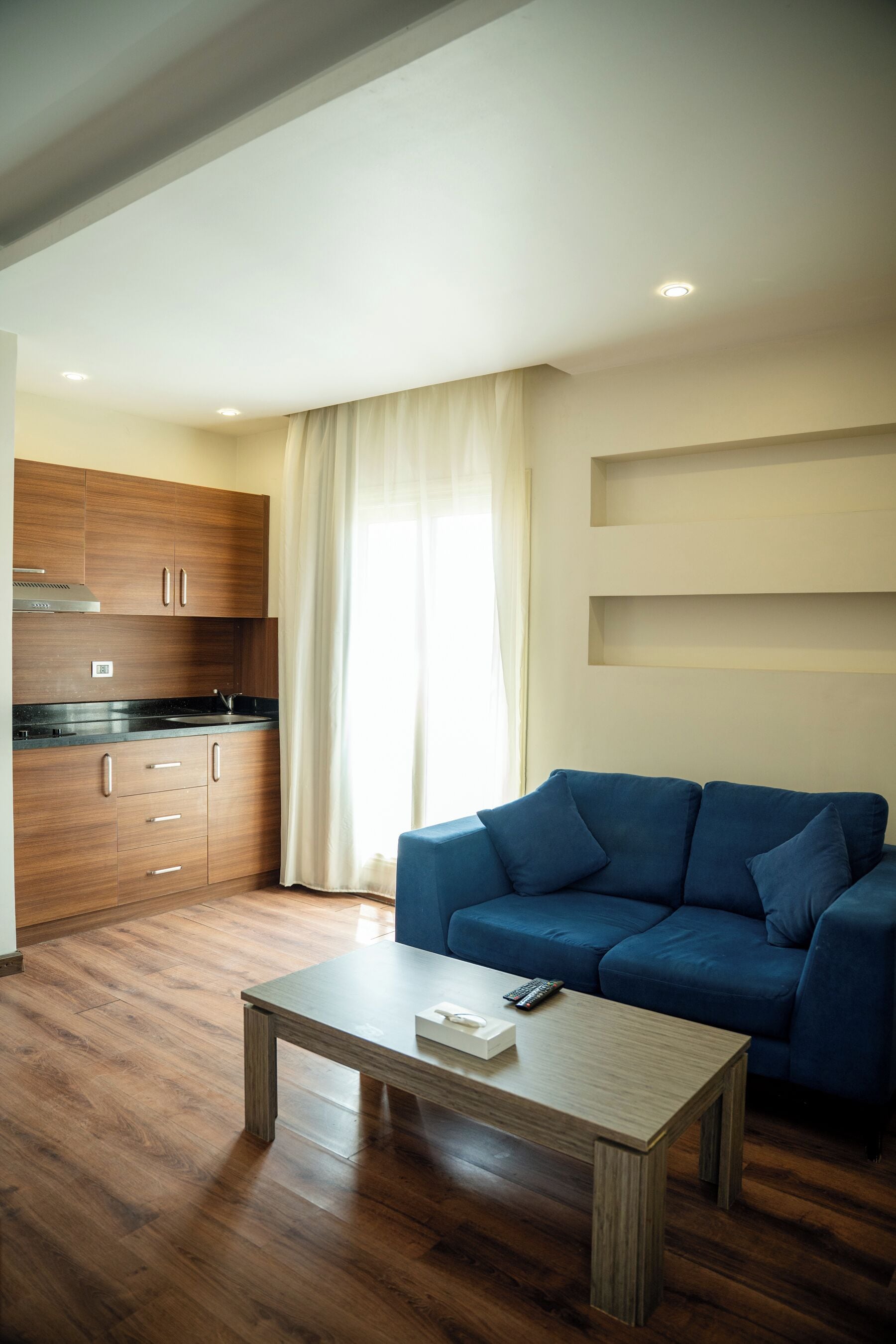 Una sala de estar moderna con un sofá azul, una mesa de centro de madera y una cocina con armarios de madera y encimera negra.