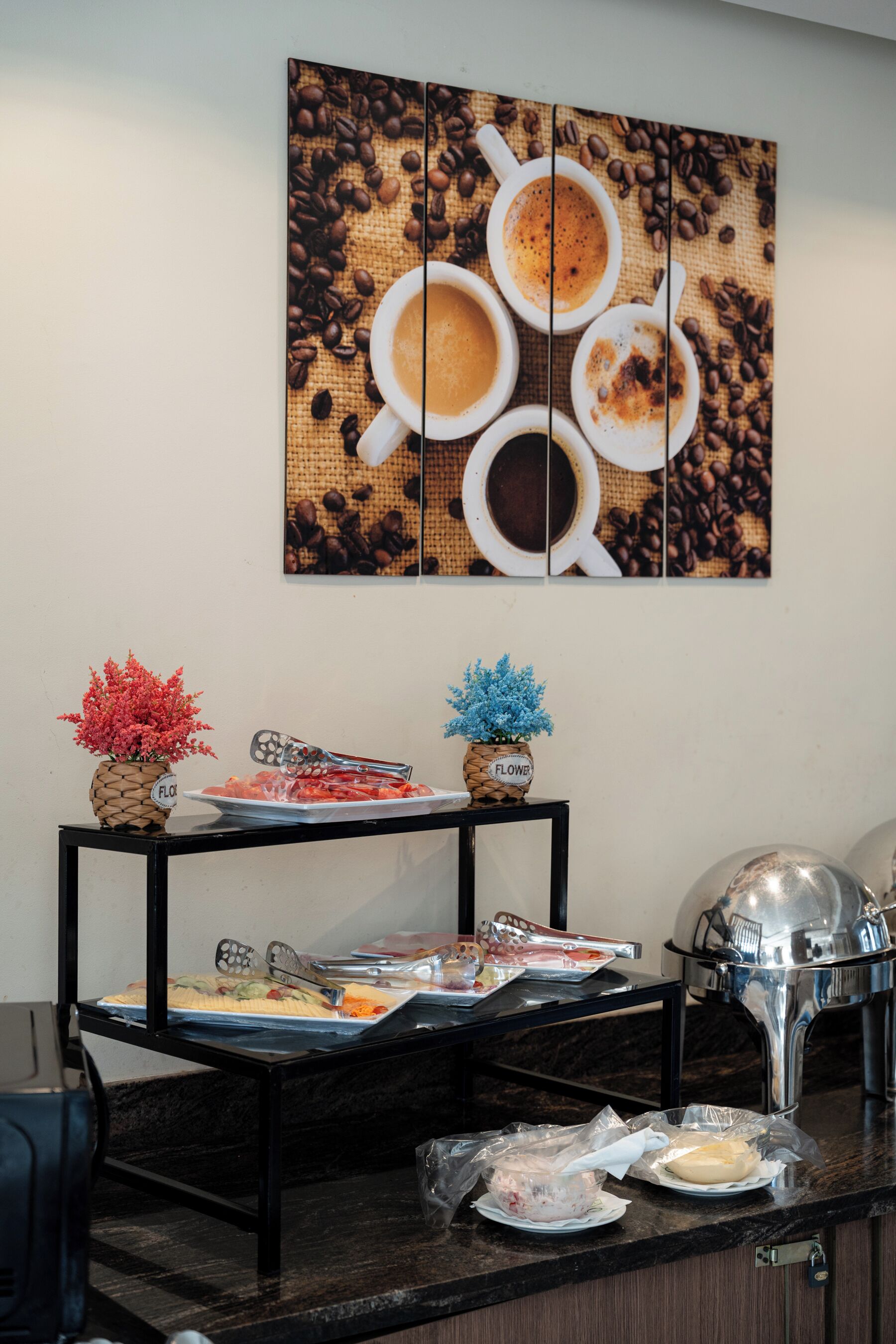 A buffet setup with a coffee-themed wall art, two flower vases, and a silver appliance on a black countertop.