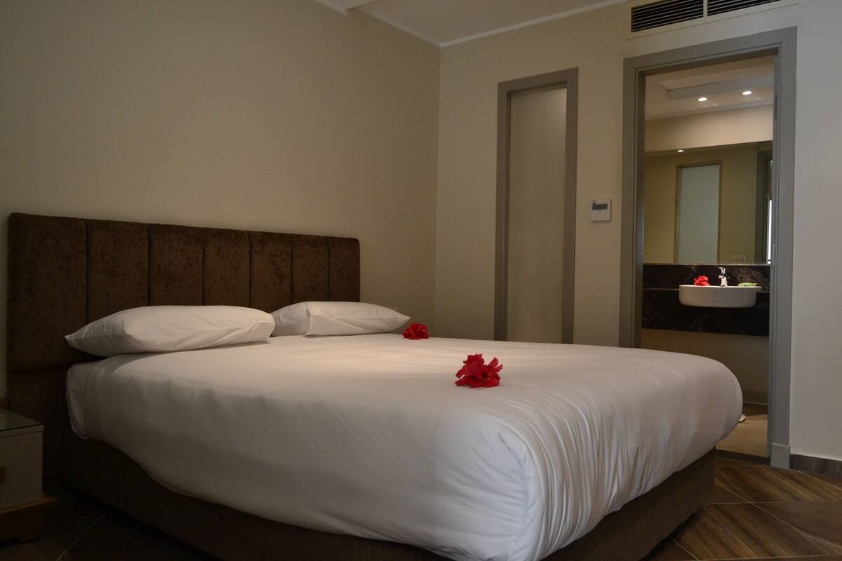 A neatly made bed with white linens and two red flowers, a wooden headboard, and a bathroom with a sink and mirror visible through an open door.