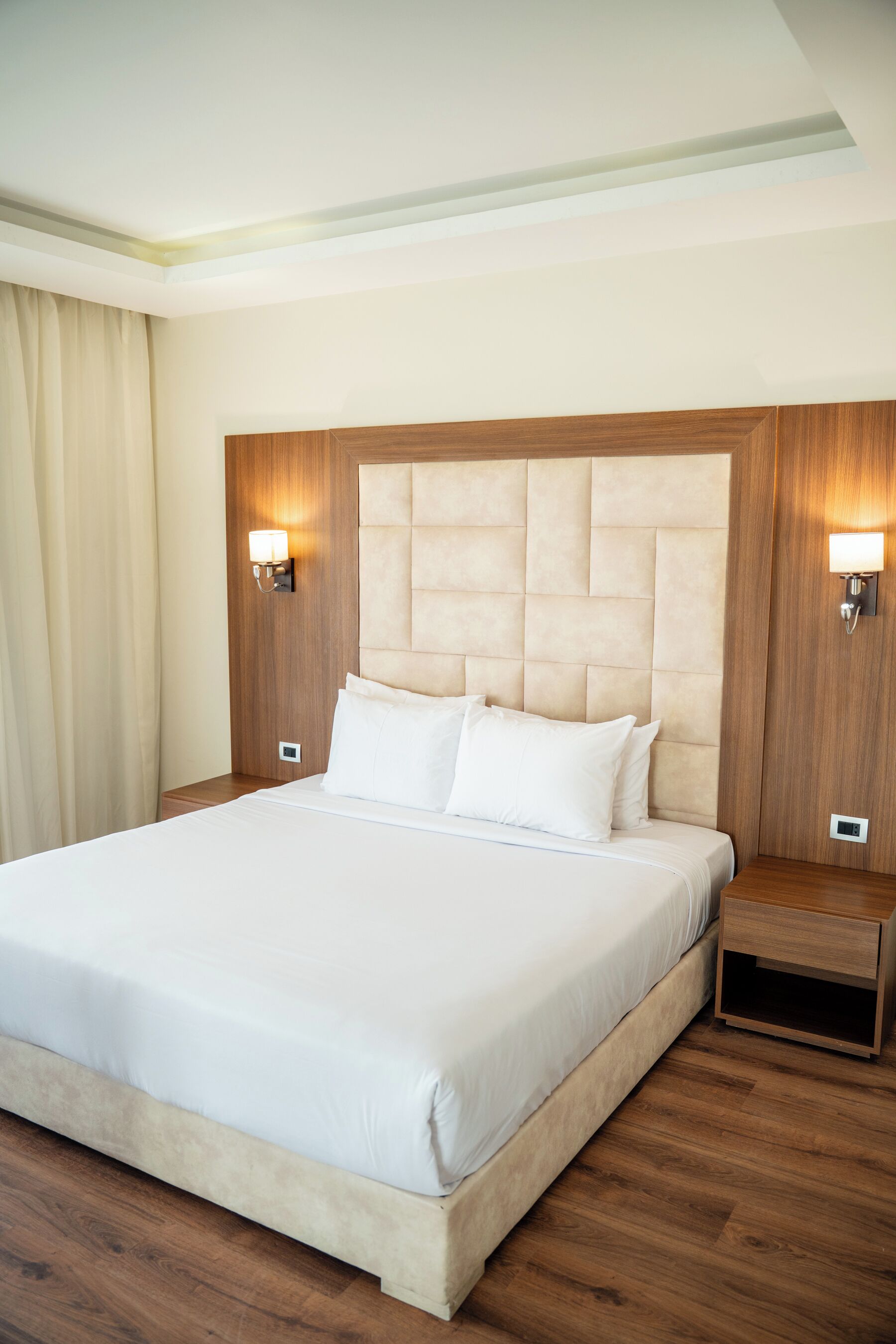Quarto de hotel com uma cama grande, cabeceira de madeira, luminárias laterais e piso de madeira.