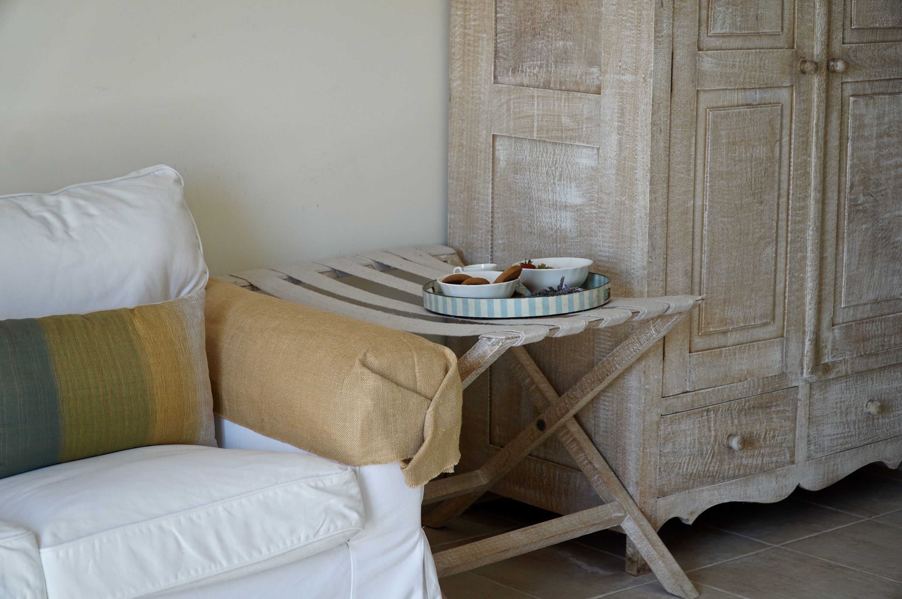 Ein weißes Sofa mit einer beige farbenen Decke, ein kleiner Tisch mit gestreiftem Tischläufer und eine Holzkommode mit Schubladen.