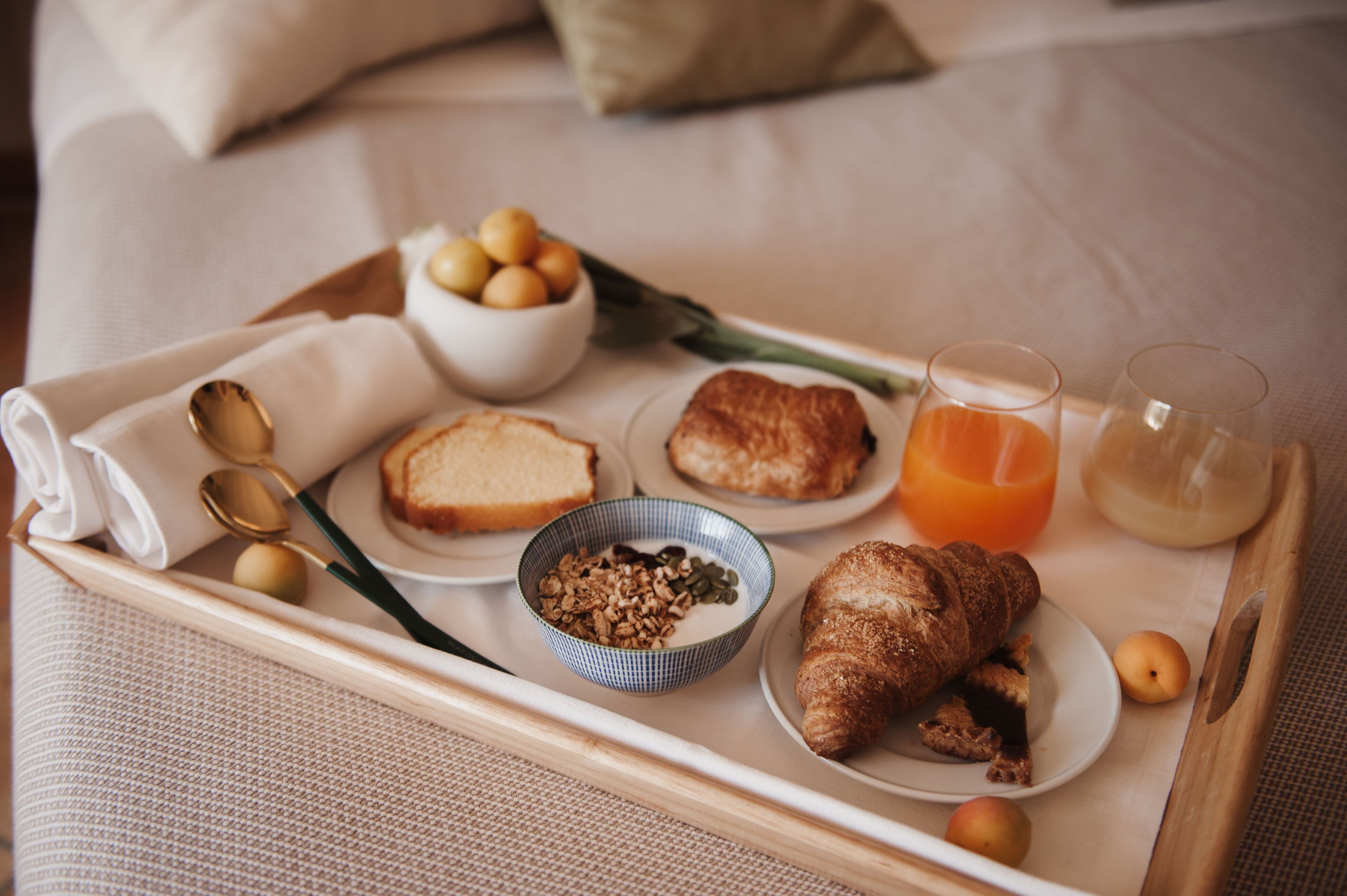 Bandeja de desayuno con tostadas, croissants, un tazón de cereal, medio croissant, un vaso de jugo de naranja y un vaso de leche sobre una cama.