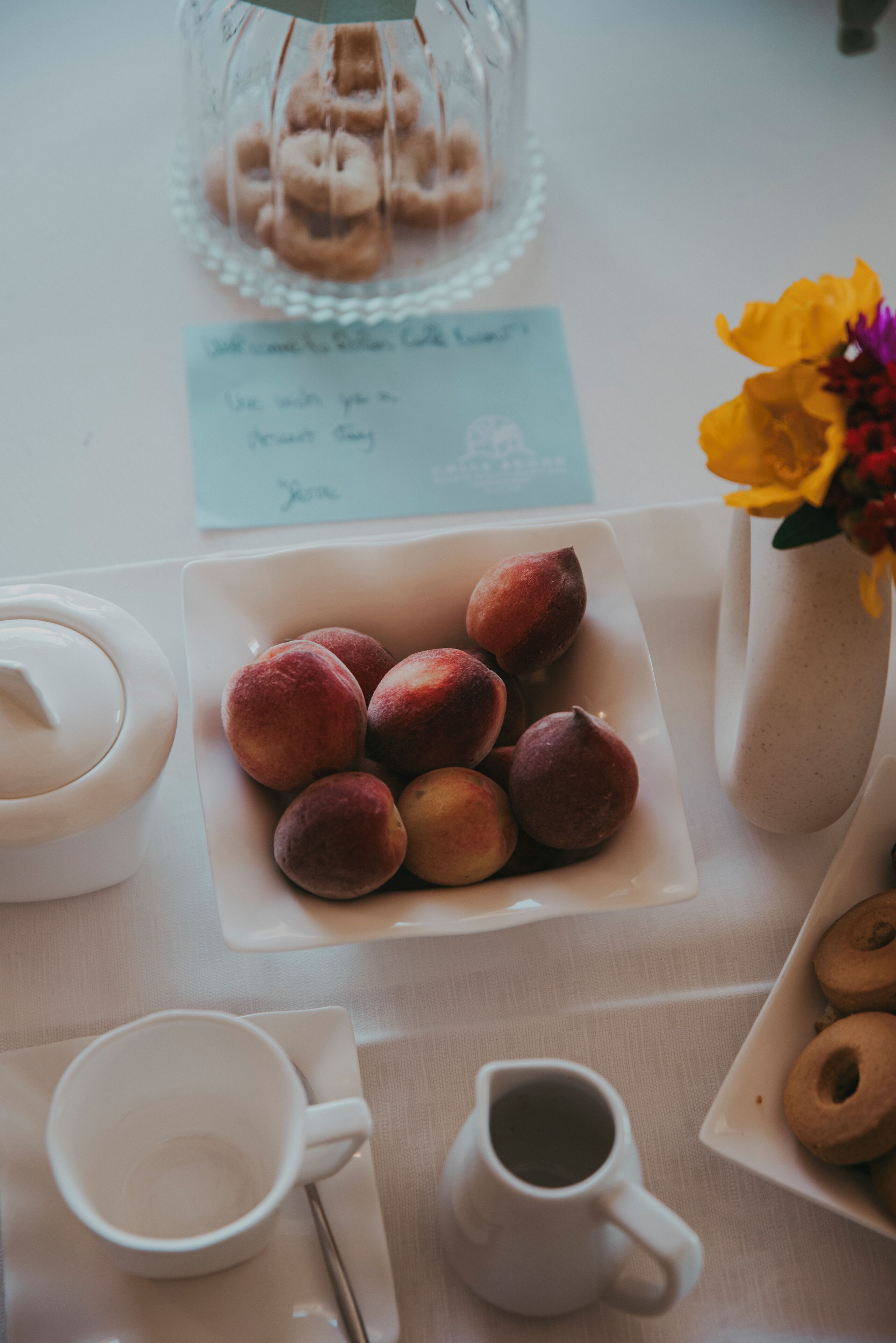A table with a plate of peaches, a pitcher of coffee, a cup, a glass jar with doughnuts, and a vase with flowers.