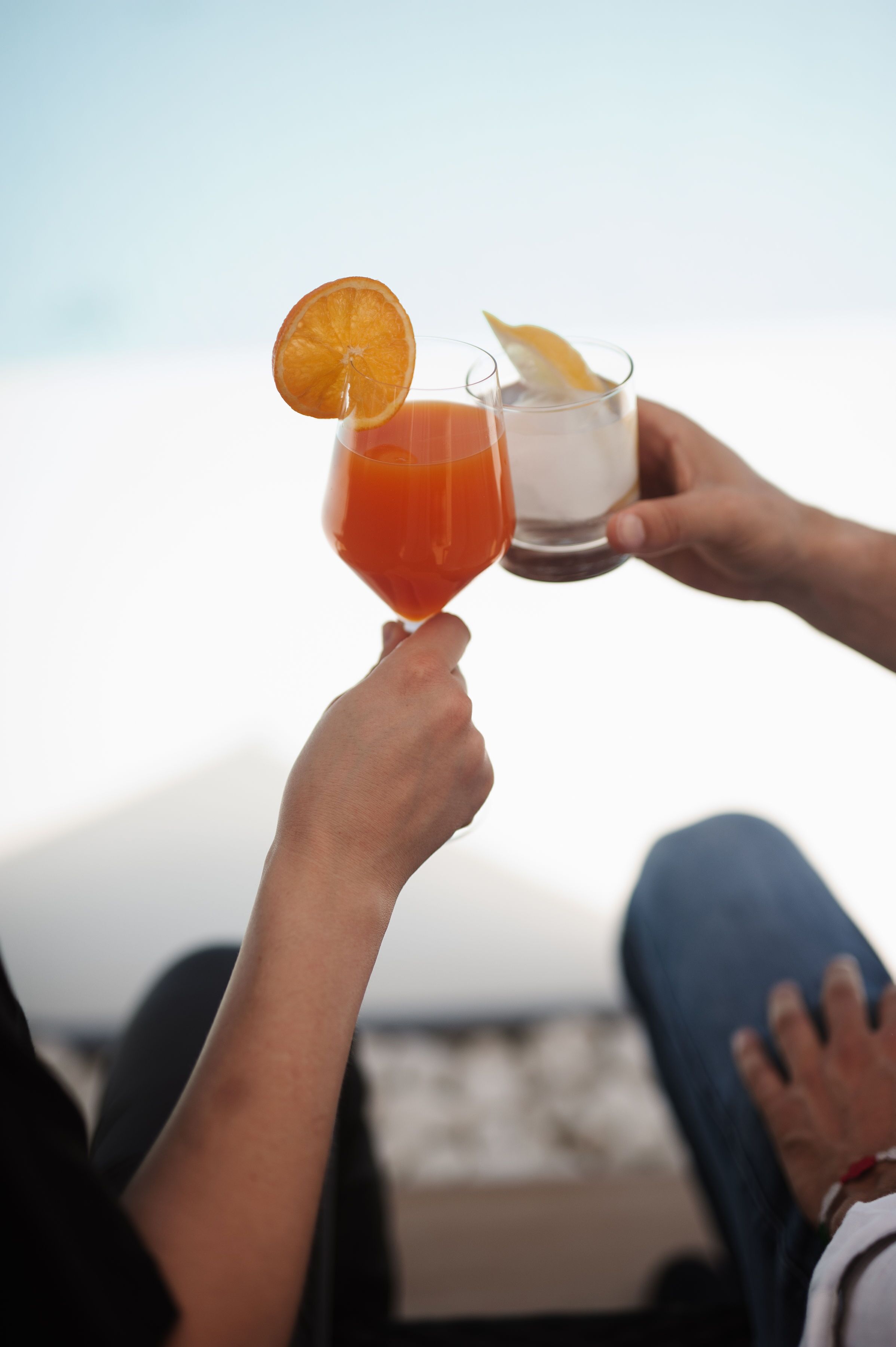Two people holding cocktail glasses with garnishes, sitting by a body of water.