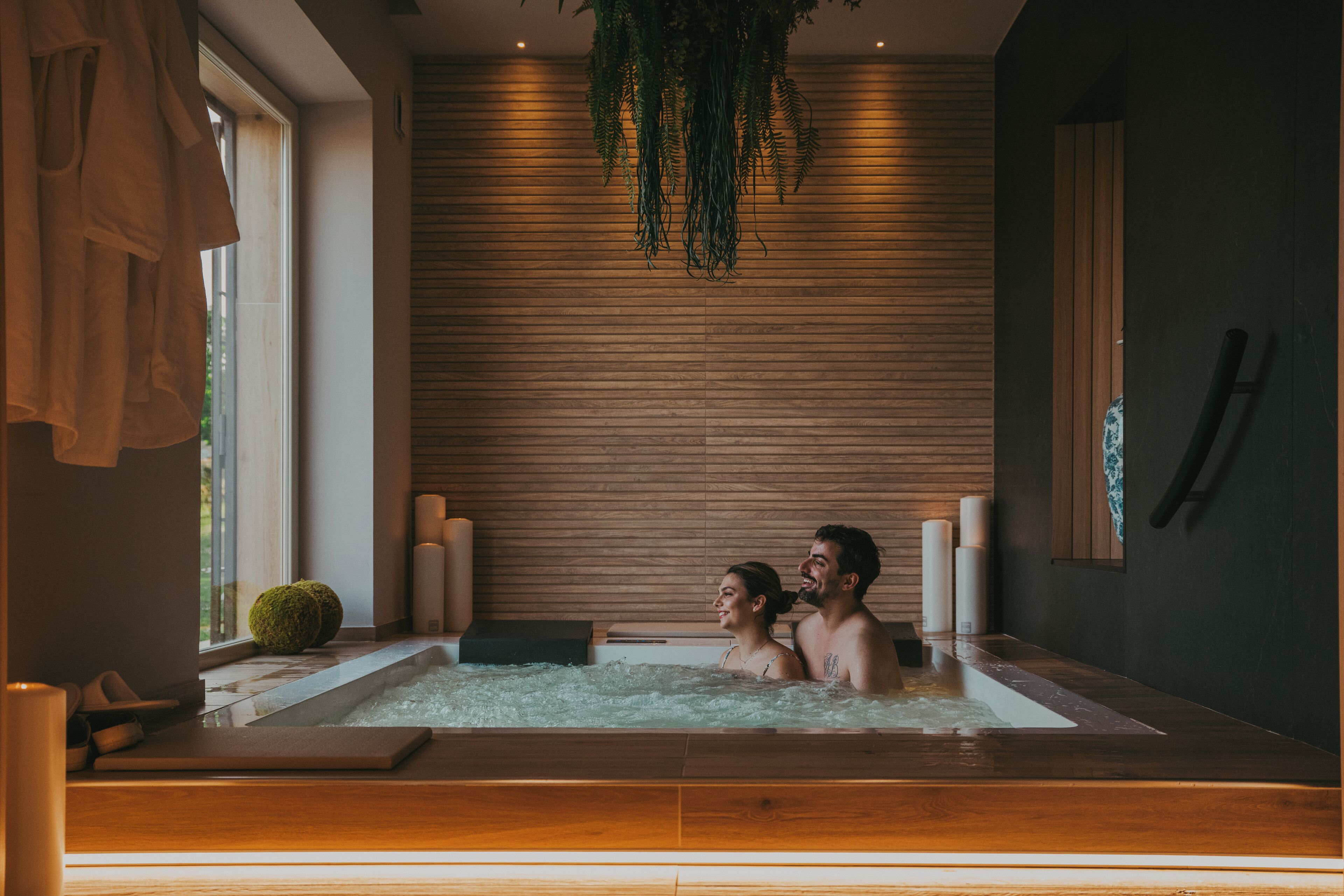 Ein Paar in einer modernen Spa-Badewanne mit Holzvertäfelung und einer hängenden Pflanze.