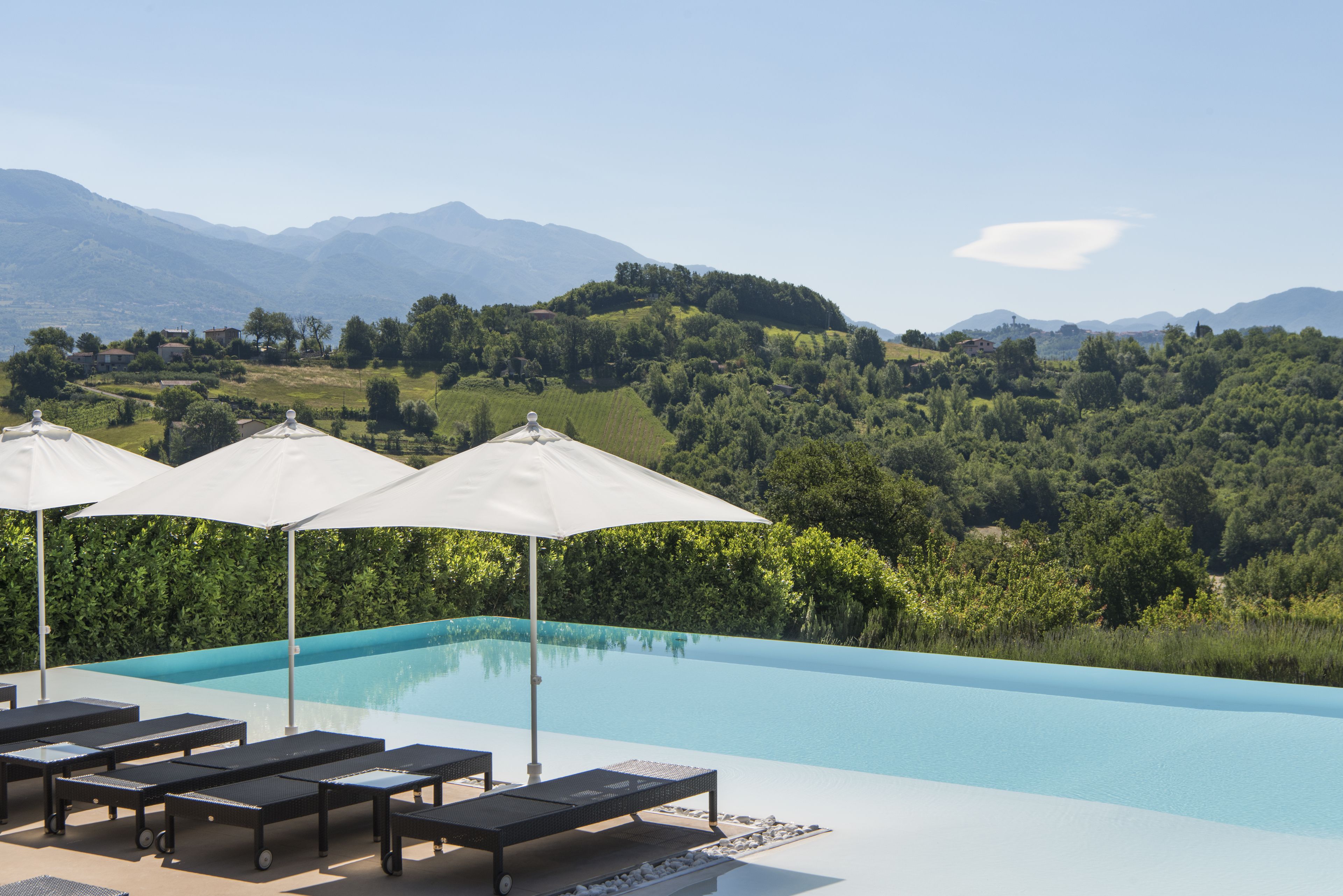 Zona de piscina con sillones de descanso y sombrillas, rodeada de exuberante vegetación y montañas al fondo.