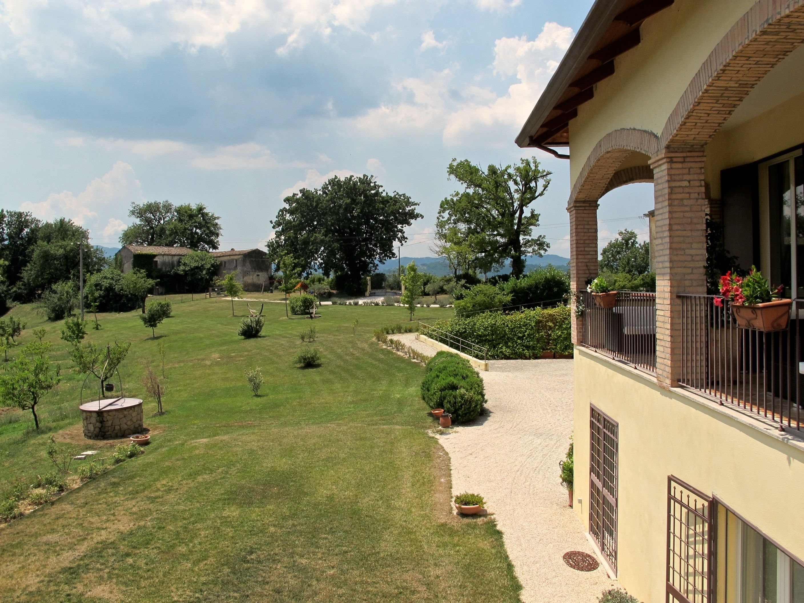 A house with a balcony, a garden with a stone well, and a view of trees and mountains.