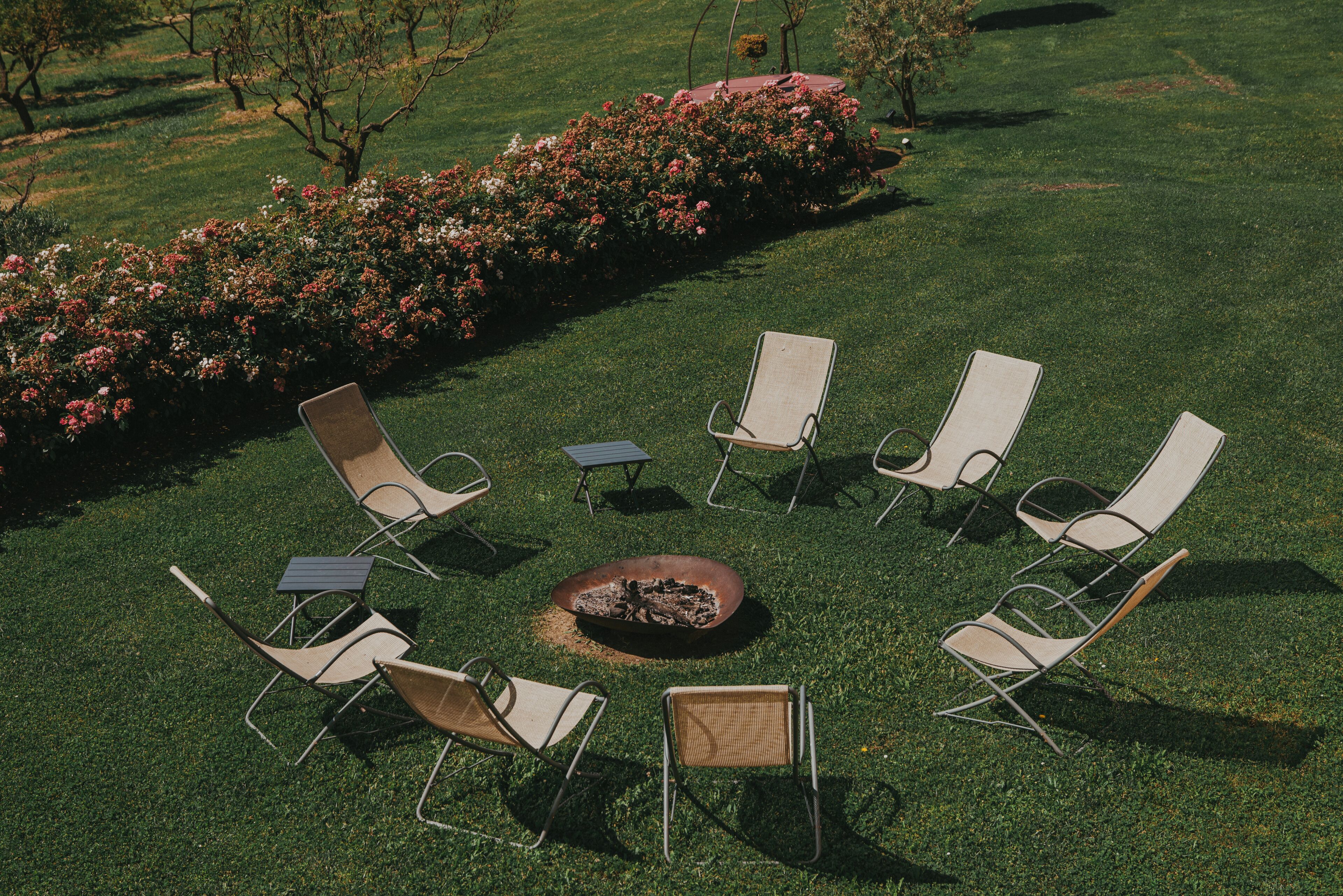 A circular arrangement of lawn chairs around a central fire pit on a well-maintained lawn with a flower bed in the background.