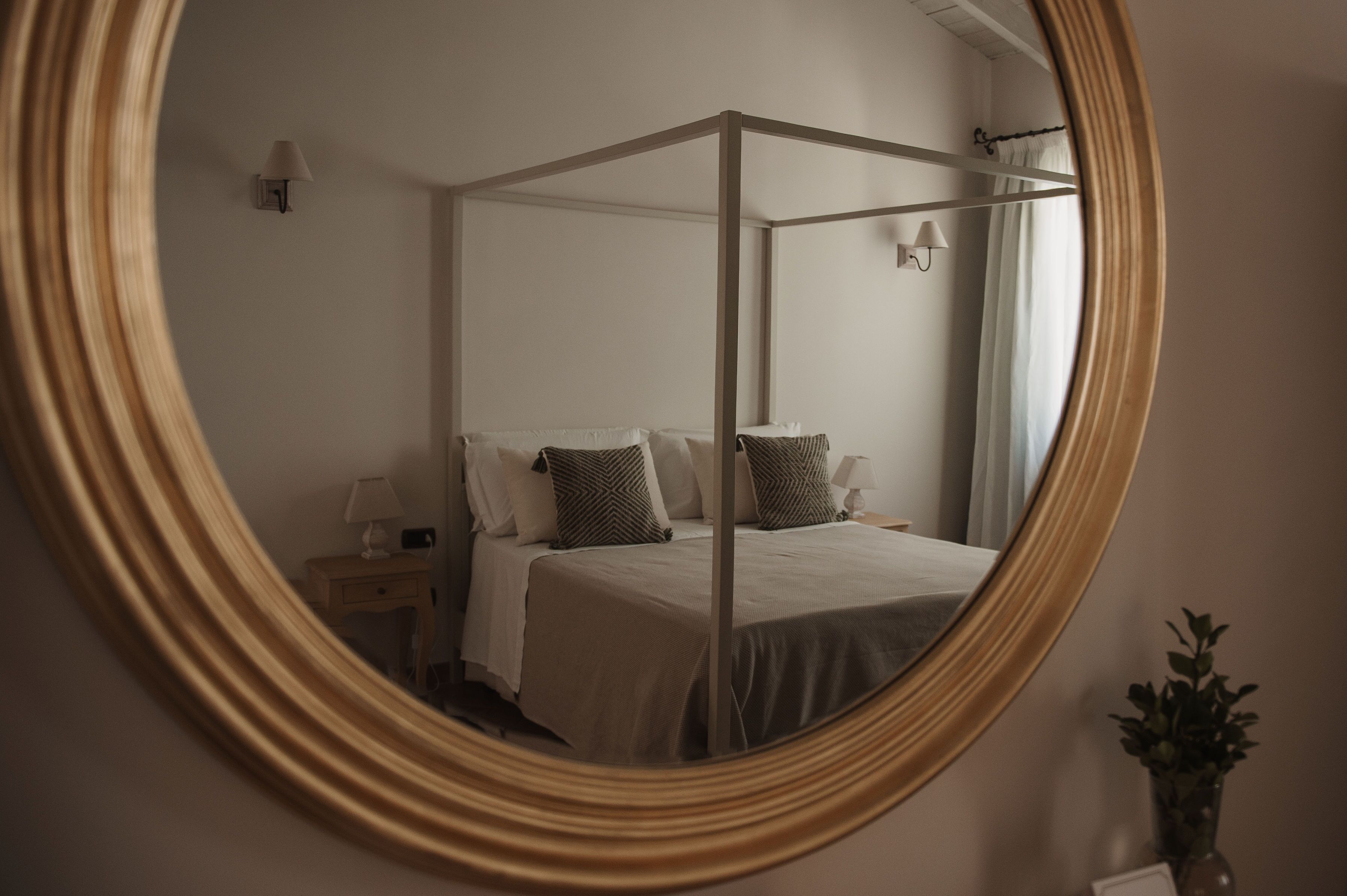 Una cama con dosel, con una colcha blanca y dos almohadas decorativas, una mesita de noche con una lámpara, y un espejo en la pared.