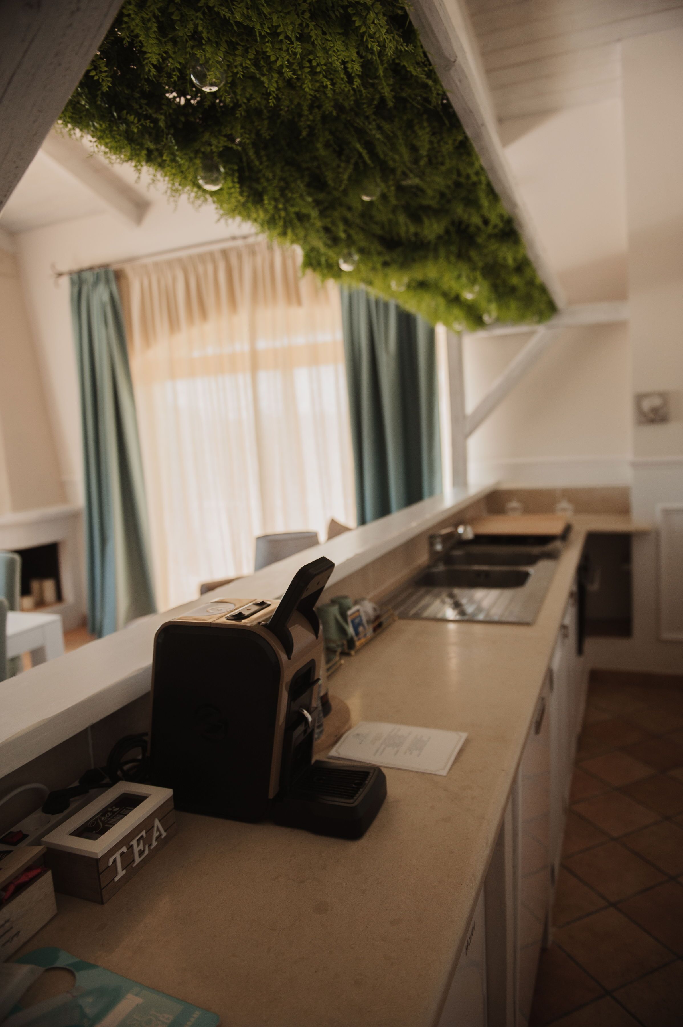 Una cocina con cafetera, caja de té y ventana con cortinas.