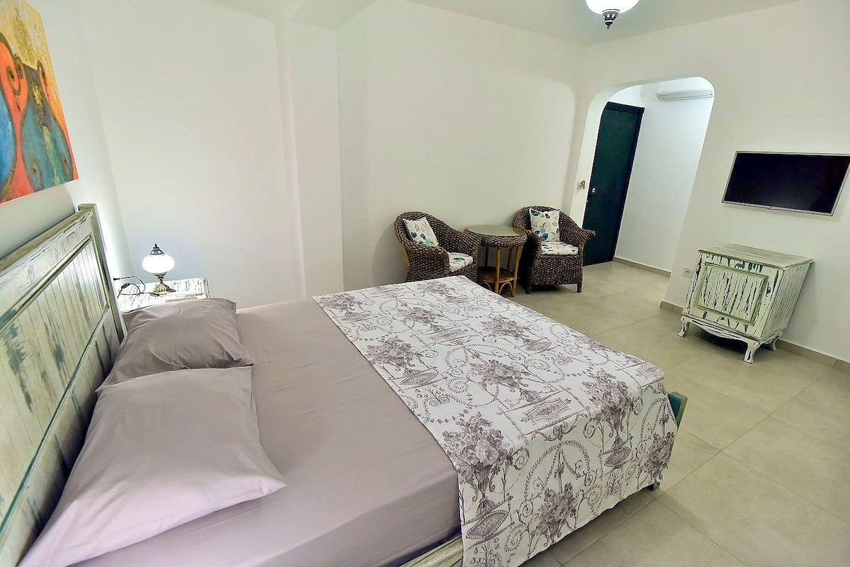 Un dormitorio con cama, una silla, una mesita, televisor y radiador.