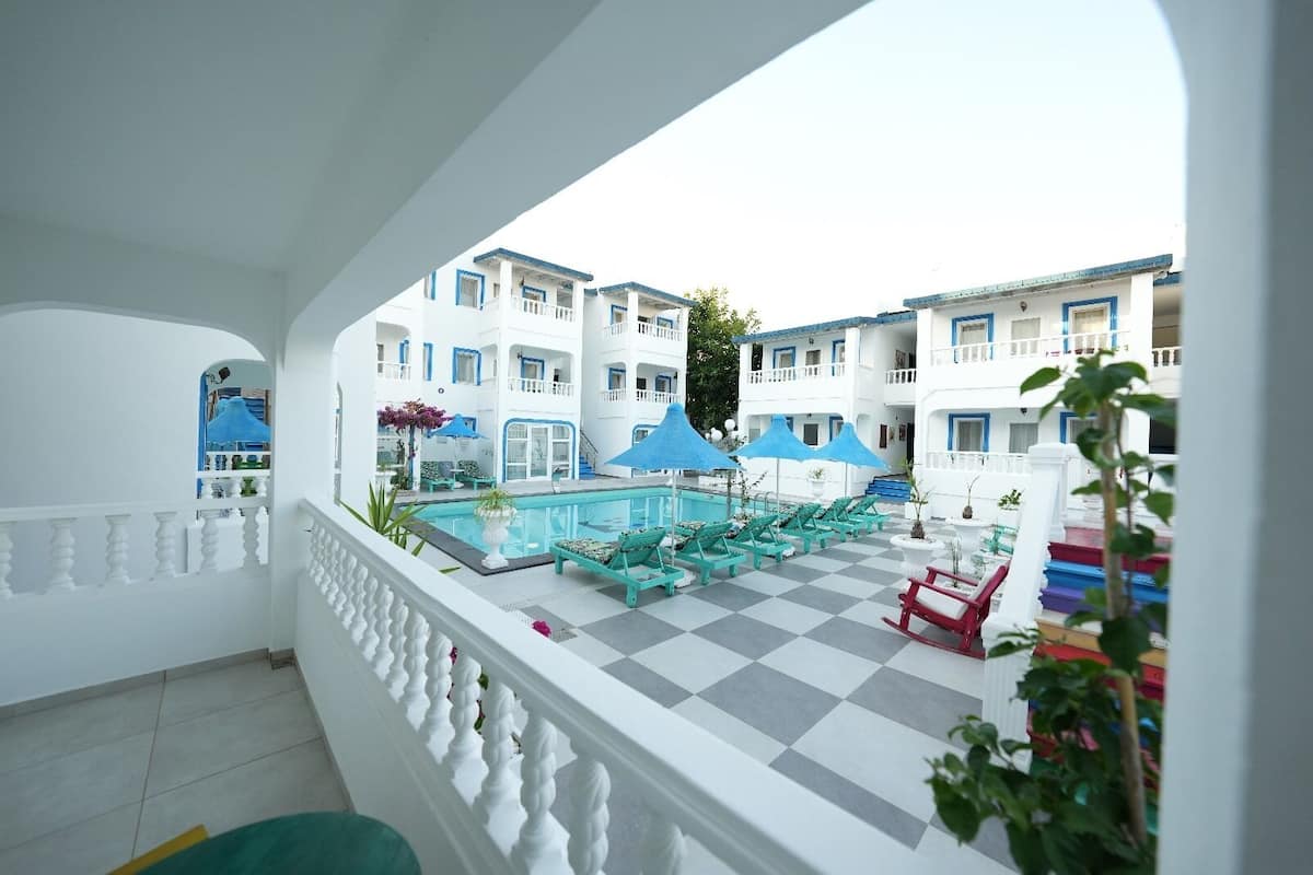 Un área de piscina con múltiples sillas de descanso y sombrillas, rodeada de edificios blancos con balcones.