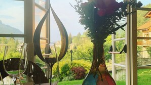A table with wine glasses and a vase with flowers, set against a window with a view of greenery and buildings. - T'AMI Hotel Resort SPA (Selvino)