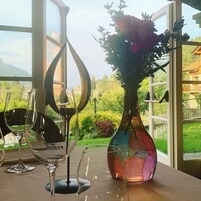 Una mesa con copas de vino y un florero, frente a una ventana con vistas a zonas verdes y edificios.
