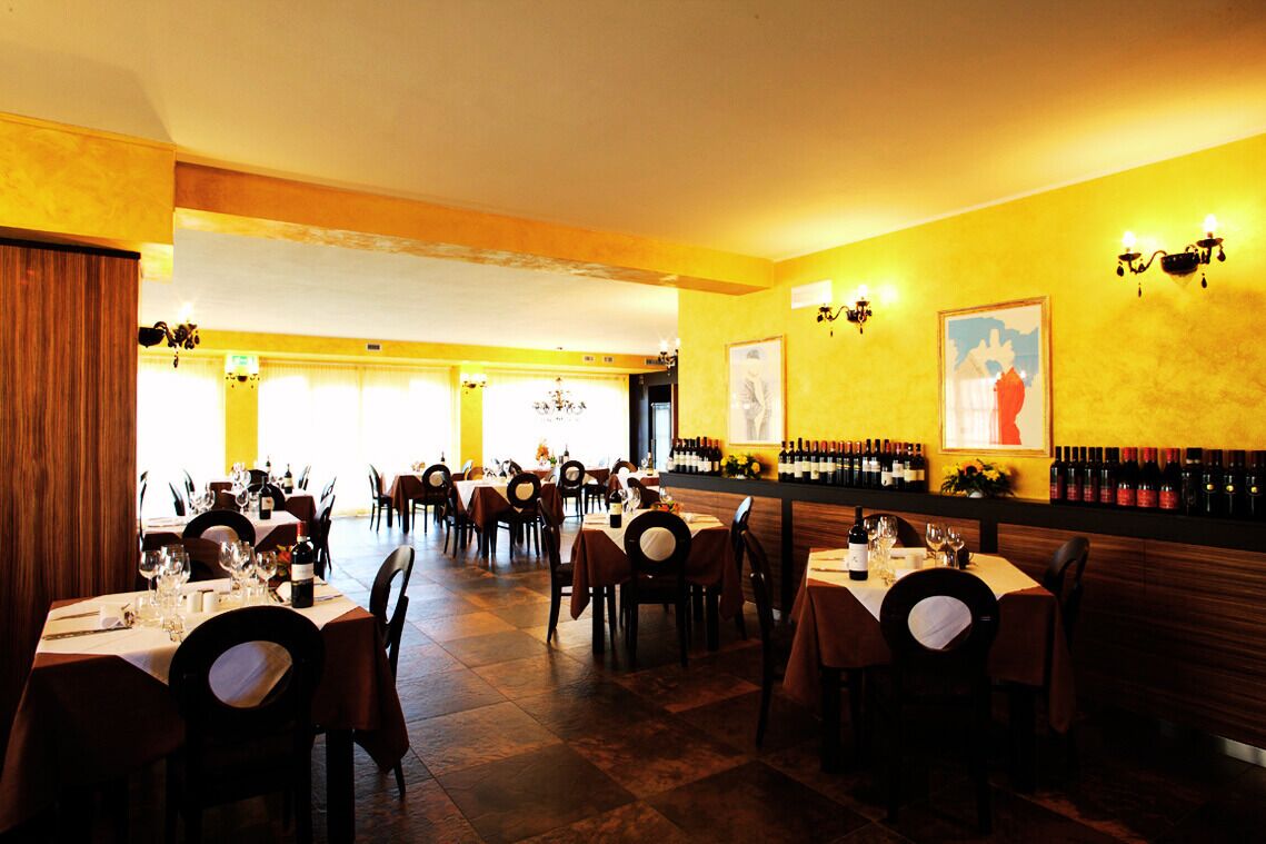 A restaurant with multiple tables set for dining, a bar area with wine bottles, and a yellow wall with framed artwork.