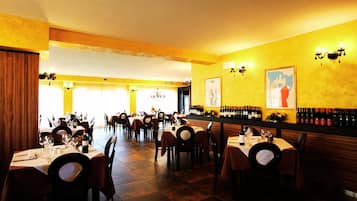 Un restaurant avec plusieurs tables dressées pour le service, un coin bar avec des bouteilles de vin et un mur jaune orné d’œuvres d’art encadrées.