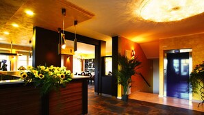 A hotel lobby with a reception desk, a bar area, and a staircase leading to an elevator. - T'AMI Hotel Resort SPA (Selvino)
