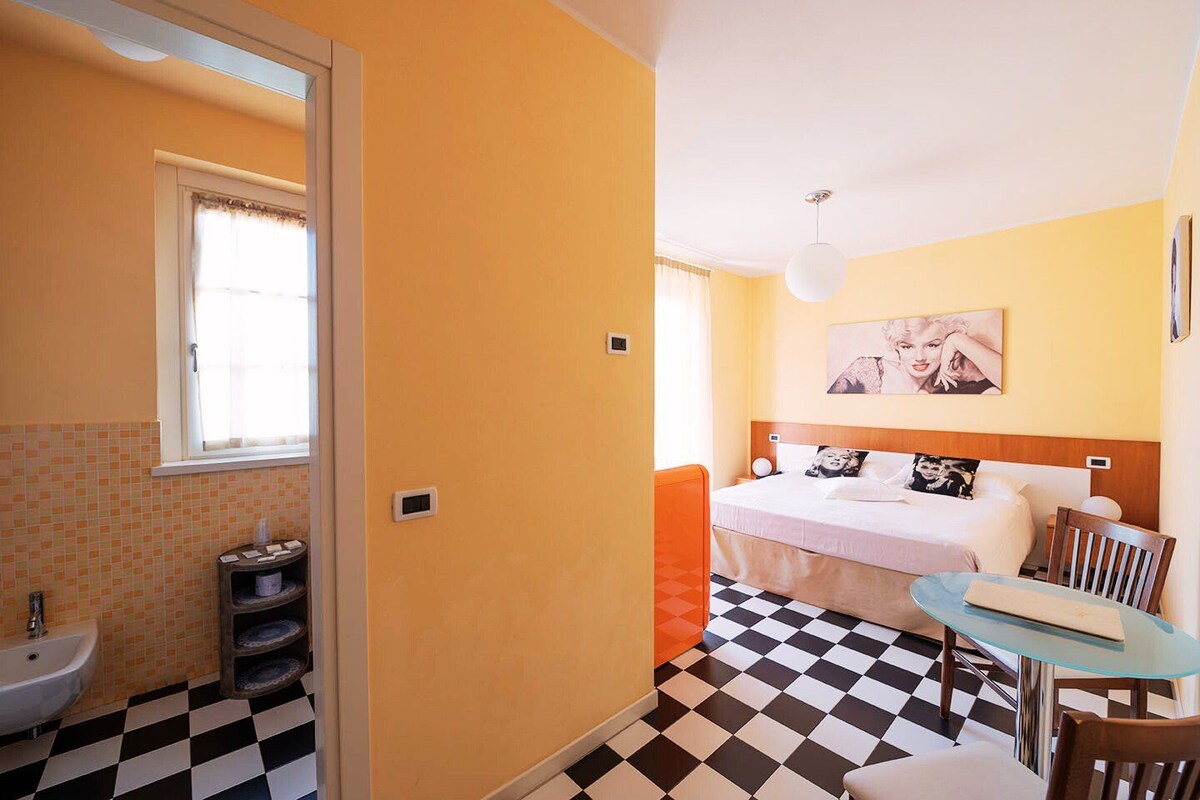 A hotel room with a checkered floor, a bed with two pillows, a bedside table with a lamp, and a window with curtains.