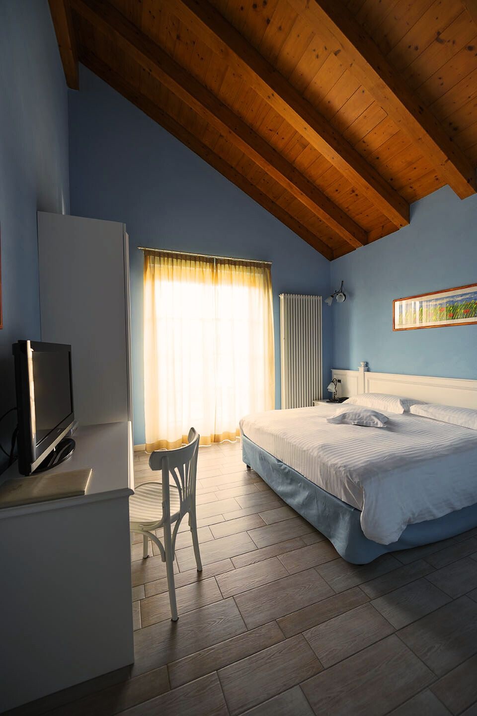A bedroom with a bed, a chair, a television, a radiator, and a window with curtains.