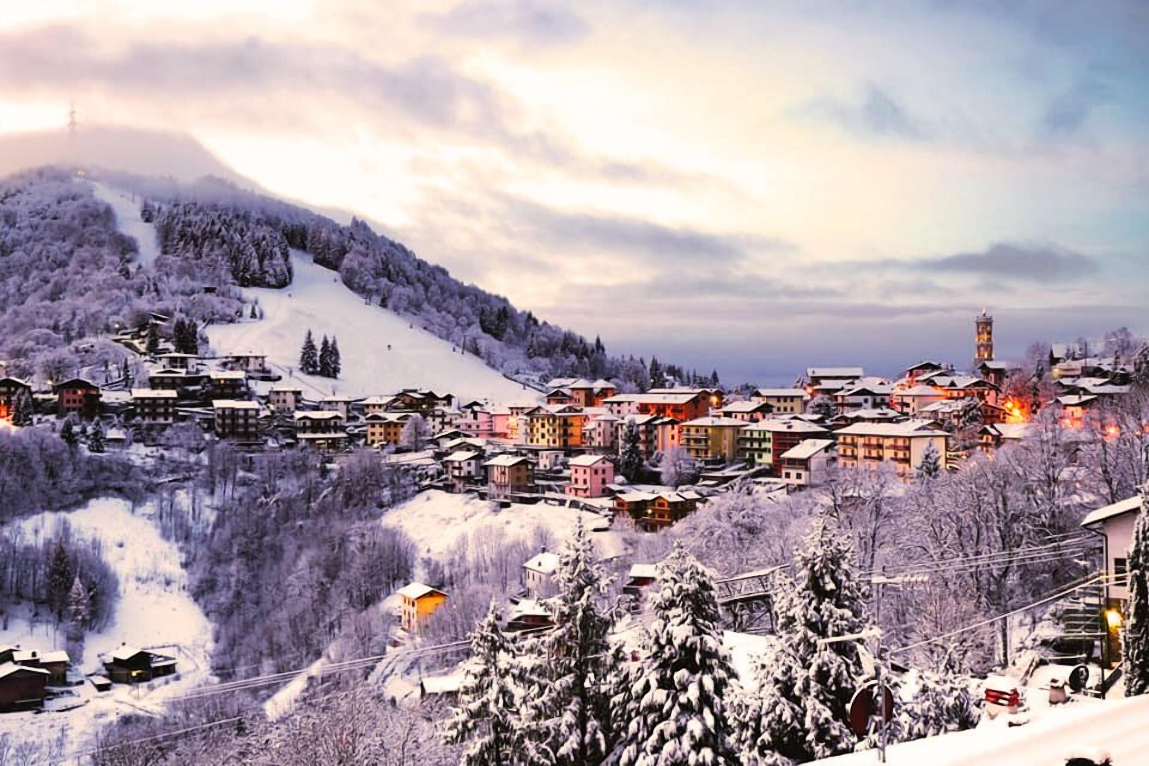 A snow-covered village with buildings and a tower, surrounded by trees and a ski slope.