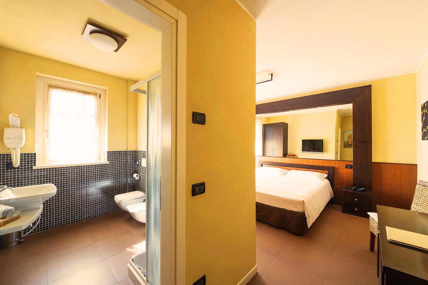Quarto de hotel com banheiro, cama, televisão e escrivaninha.