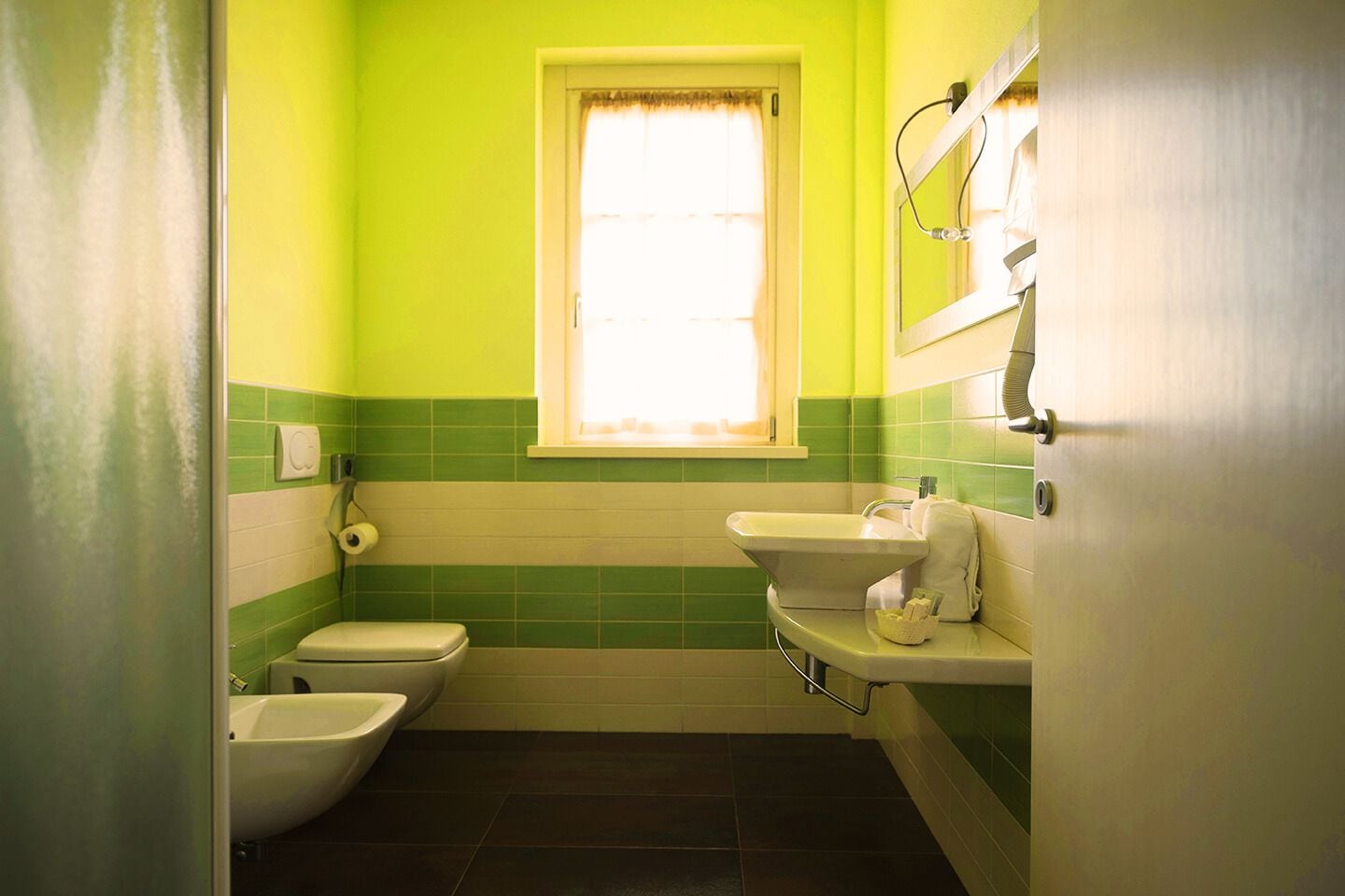Un baño pequeño con inodoro, bidé y lavamanos, paredes verdes y una ventana con cortinas.