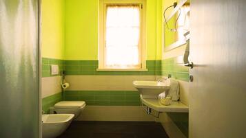 Un baño pequeño con inodoro, bidé y lavamanos, paredes verdes y una ventana con cortinas.