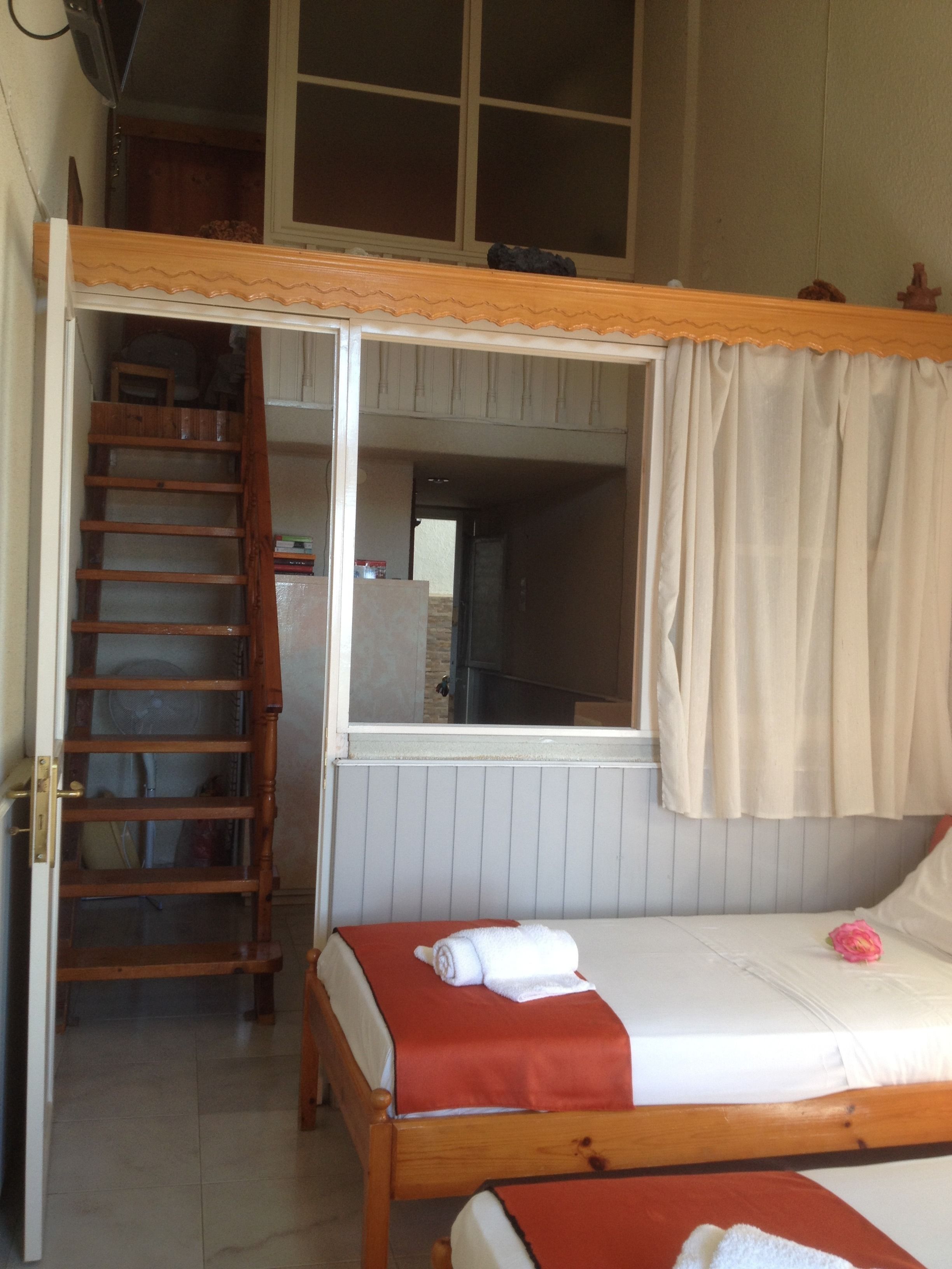 A dorm-style room with a loft bed, a wooden ladder, a window with curtains, and a small kitchenette.