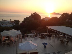 Une terrasse avec des sièges extérieurs, une table avec un parasol et une vue sur le coucher de soleil sur la mer.