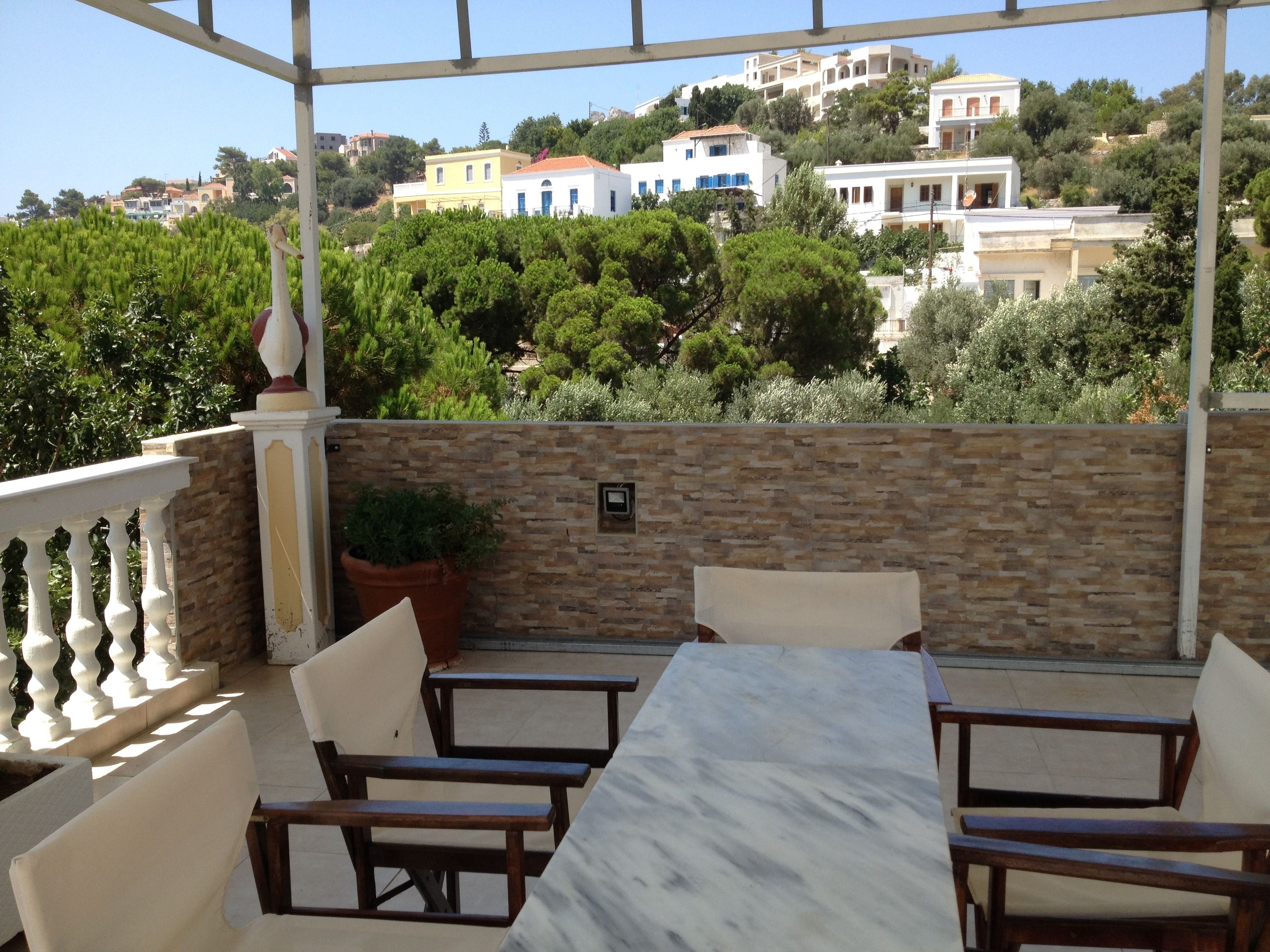 A terrace with a marble table and wooden chairs, overlooking a hillside with buildings and greenery.