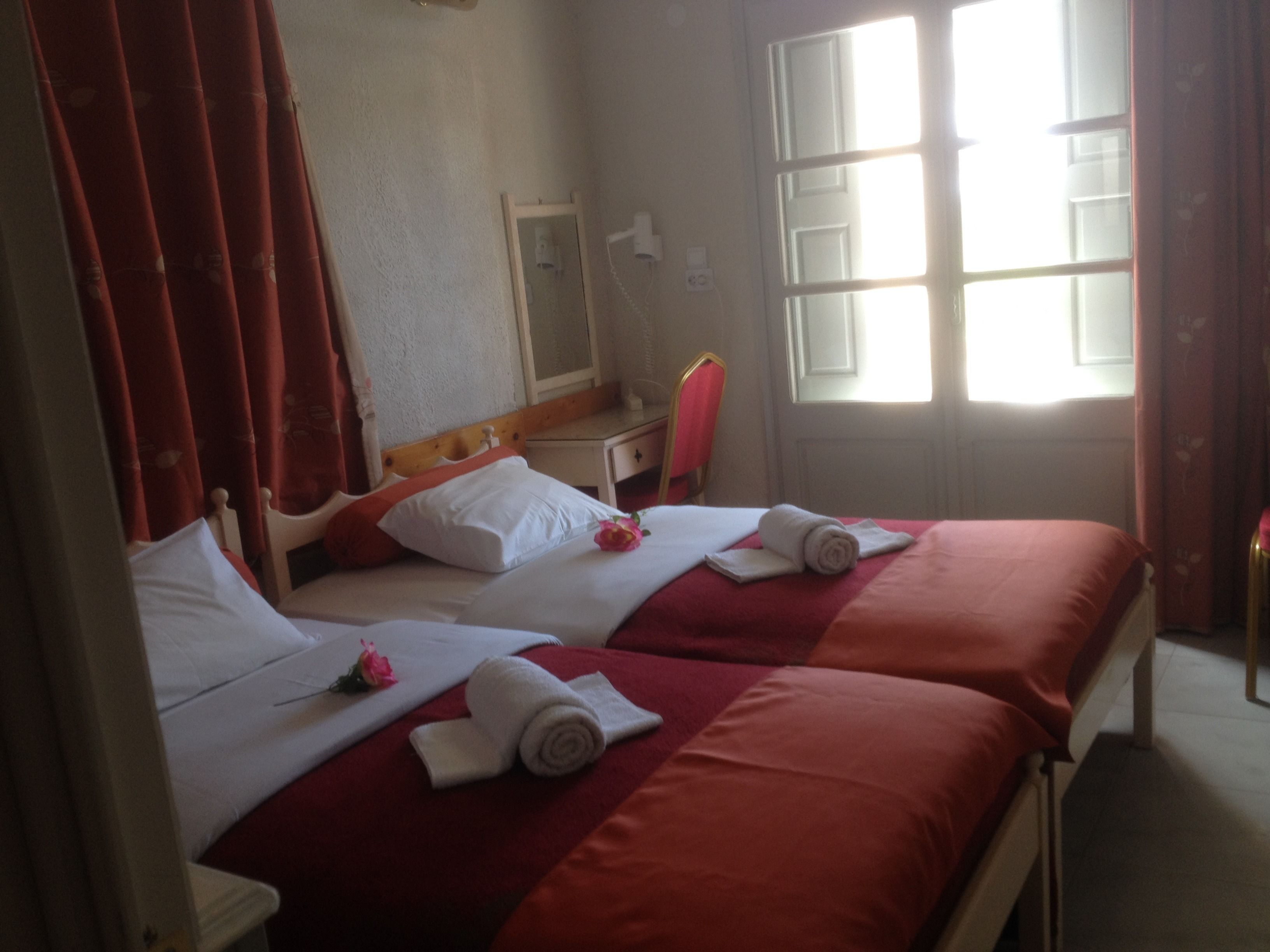 Quarto de hotel com duas camas, roupa de cama vermelha, almofadas brancas e uma janela com cortinas.