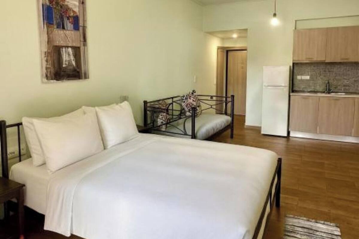 A bedroom with a bed, a nightstand, a wall-mounted painting, a kitchen area with cabinets and a refrigerator, and a wooden floor.