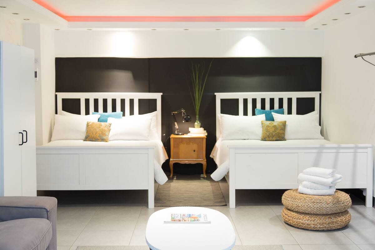 A modern hotel room with two beds, a small wooden side table, a round coffee table, and a grey sofa.