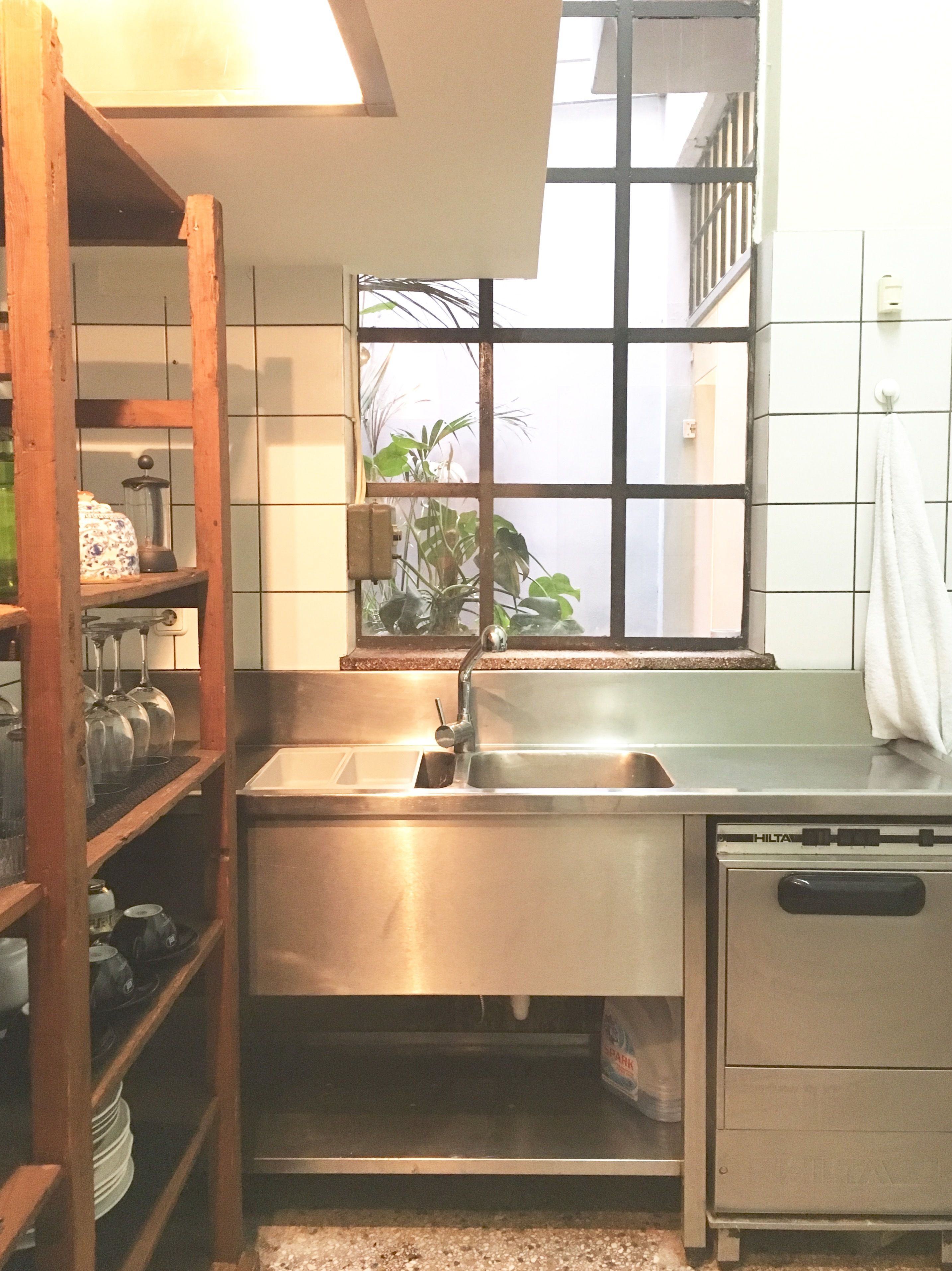 A kitchen with a stainless steel sink, wooden shelves, a window with a view of plants, and a tiled backsplash.
