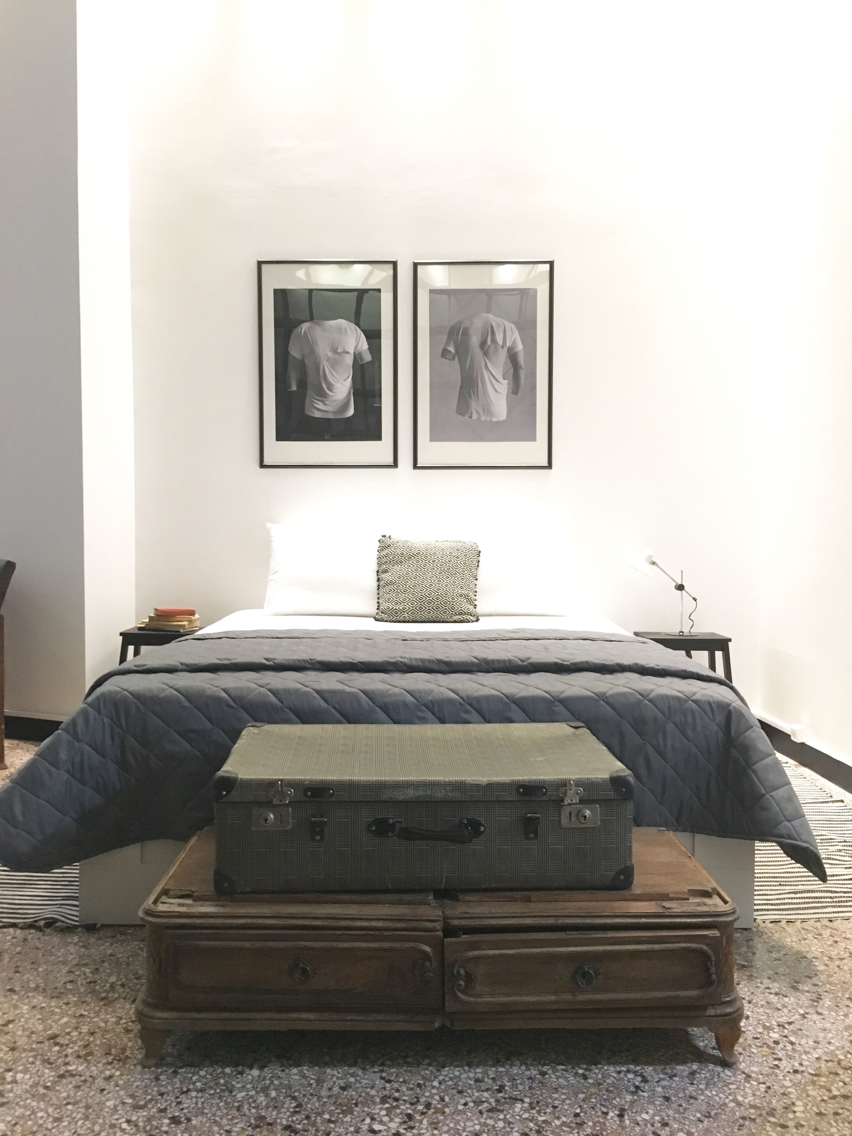 A bedroom with a bed, two framed pictures on the wall, a bedside table with a lamp, and a suitcase on a wooden chest.