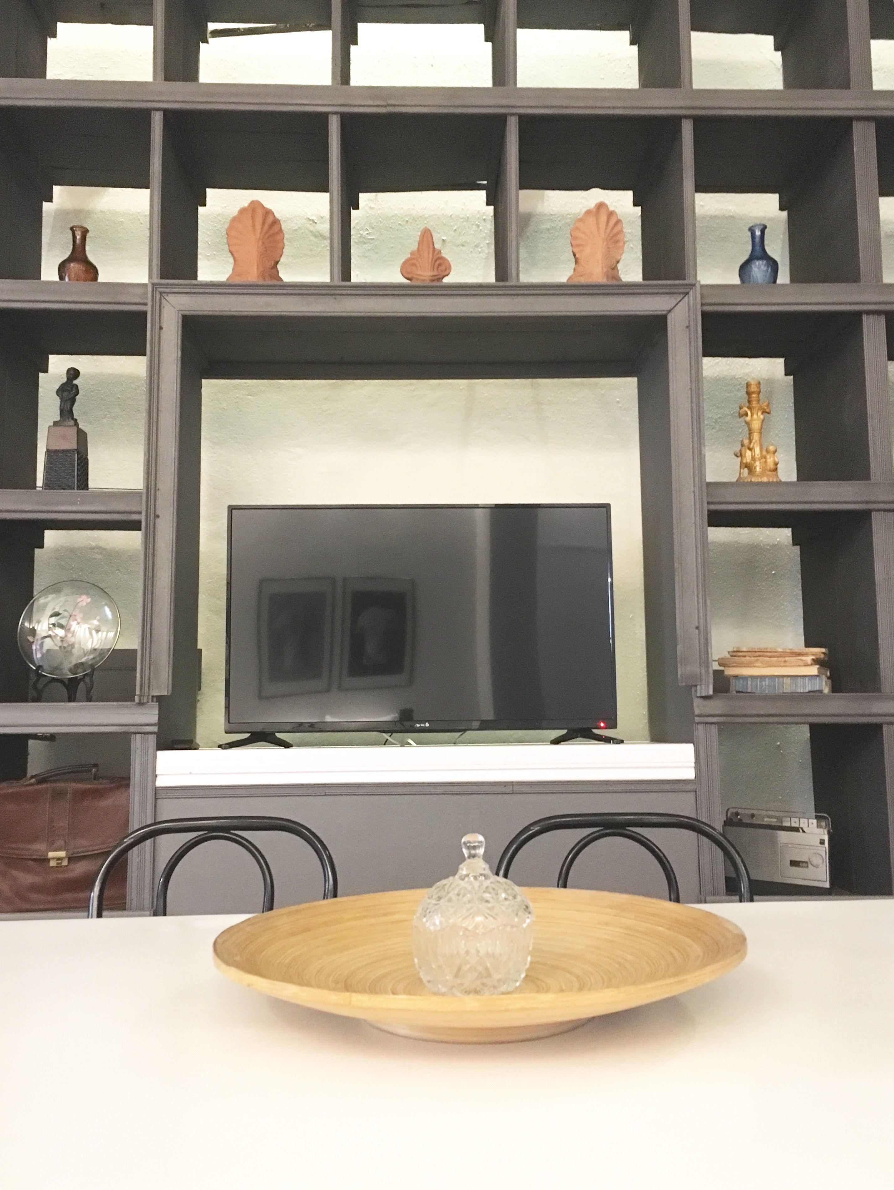 A modern living room with a flat-screen TV, wooden shelves with decorative items, and a dining table with a glass bowl centerpiece.