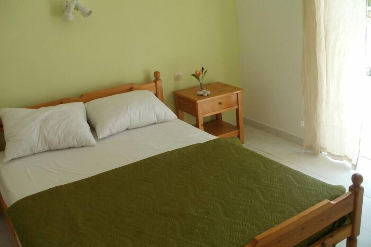 A bed with a green blanket, white pillows, and a wooden headboard. There is a small wooden nightstand with a vase and a lamp.