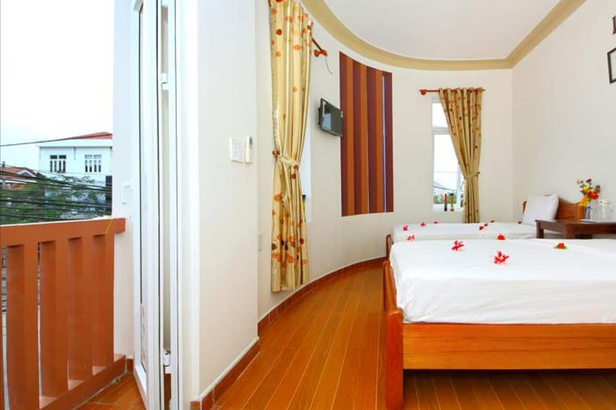 A hotel room with a wooden floor, a bed with white linens, a wooden nightstand, a small table with a vase of flowers, and a window with curtains.