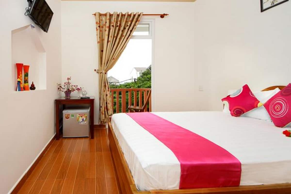 A bedroom with a bed, a small table with a refrigerator, a TV mounted on the wall, and a balcony with a wooden railing.