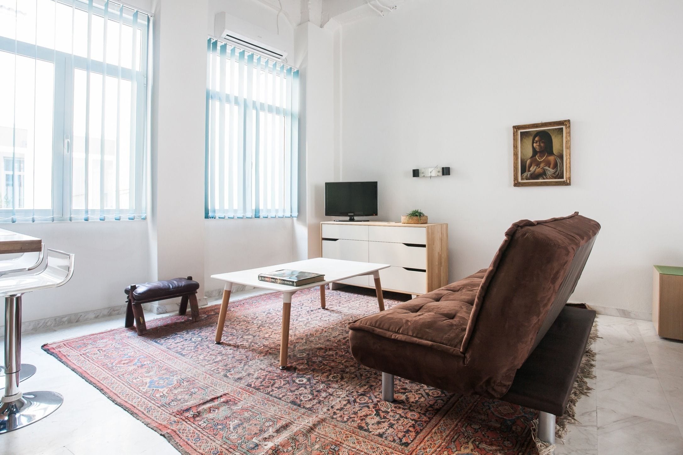 A living room with a brown sofa, a white coffee table, a TV on a wooden cabinet, a patterned rug, and a bench.