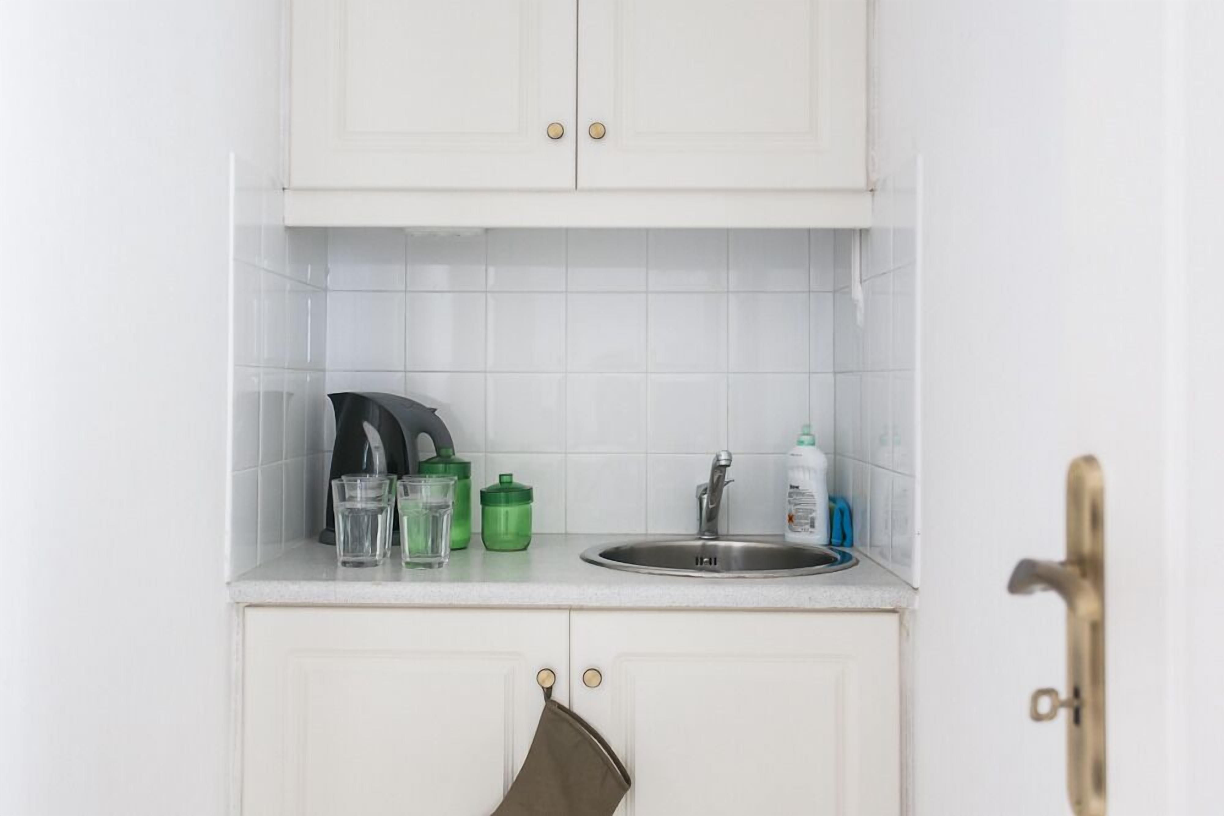 A small kitchen with white cabinets, a stainless steel sink, and a countertop with a kettle, glasses, and a green container.