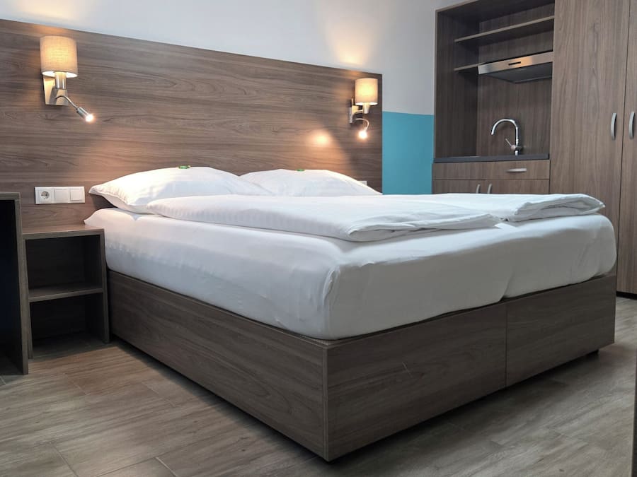 A modern hotel room with a wooden headboard, a bed with white linens, a nightstand, a desk, and a wardrobe with a mirror.